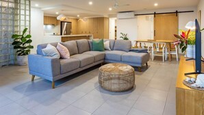 A modern living room with a grey sectional sofa, a round wicker ottoman, and a dining area with a wooden table and chairs. - Unit 4 Sundeck Gardens (Sunshine Coast)