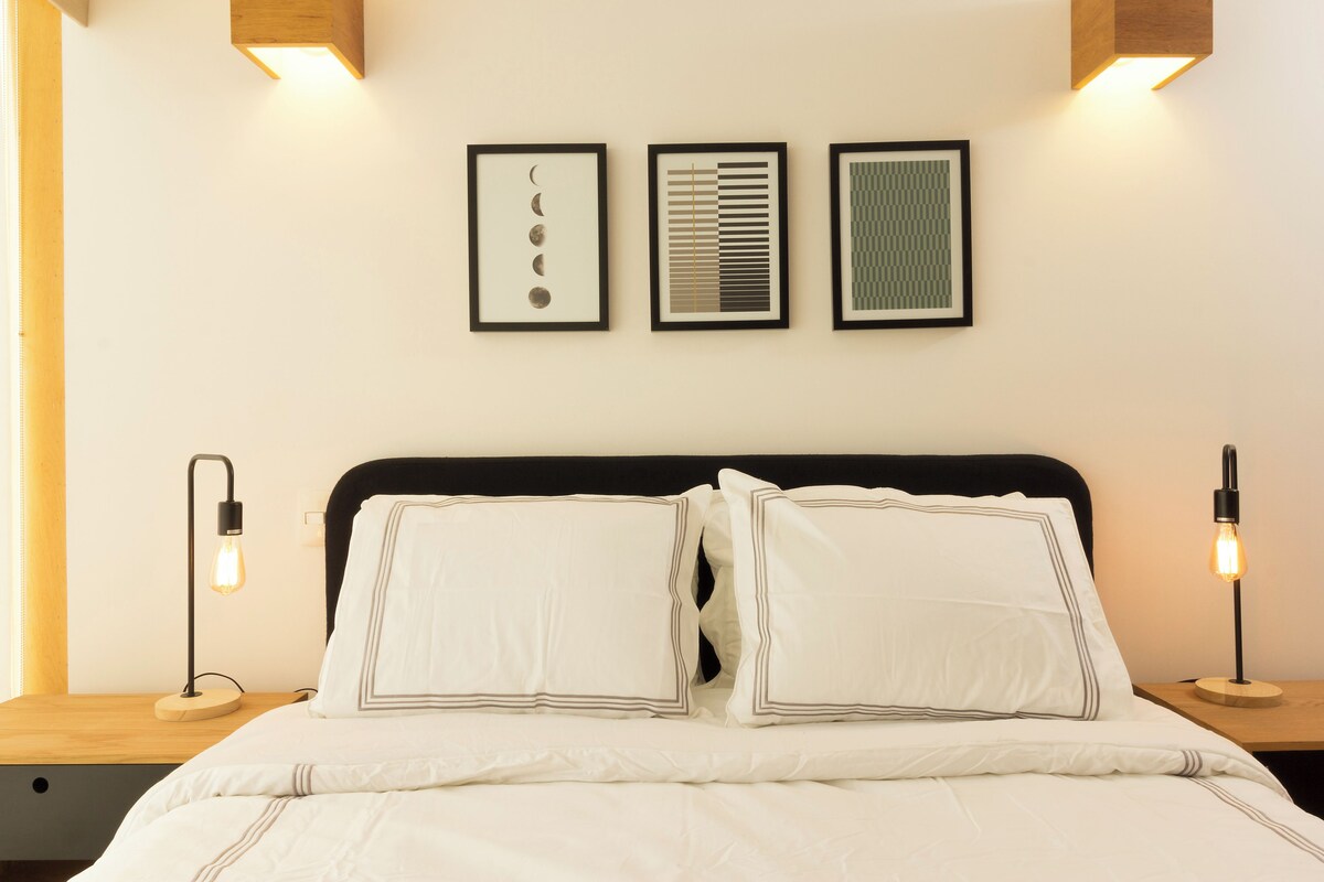 A neatly made bed with white linens and two pillows, flanked by bedside tables with lamps, and three framed artworks on the wall above.