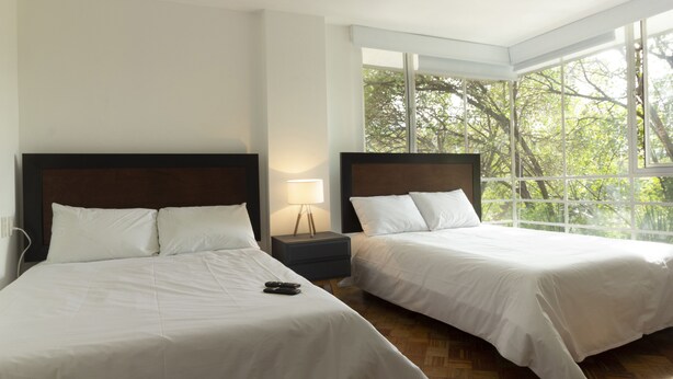 A hotel room with two beds, a nightstand, and a window with a view of trees. - Balsas By Homirent (Mexico City)