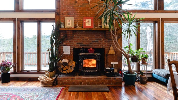 Un salón amplio con chimenea de ladrillo, vigas de madera, ventanas grandes y una variedad de plantas.