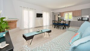 A modern living room with a sofa, a coffee table, and a dining area with chairs and a table. There is a fireplace, a TV mounted on the wall, and a kitchen area with cabinets and appliances. - A contemporary inn situated in the center of Ermioni (Ermioni)