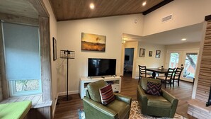 Ein geräumiges Wohnzimmer mit einem Flachbildfernseher, grünen Ledersofas, einem Essbereich und einem großen Fenster.