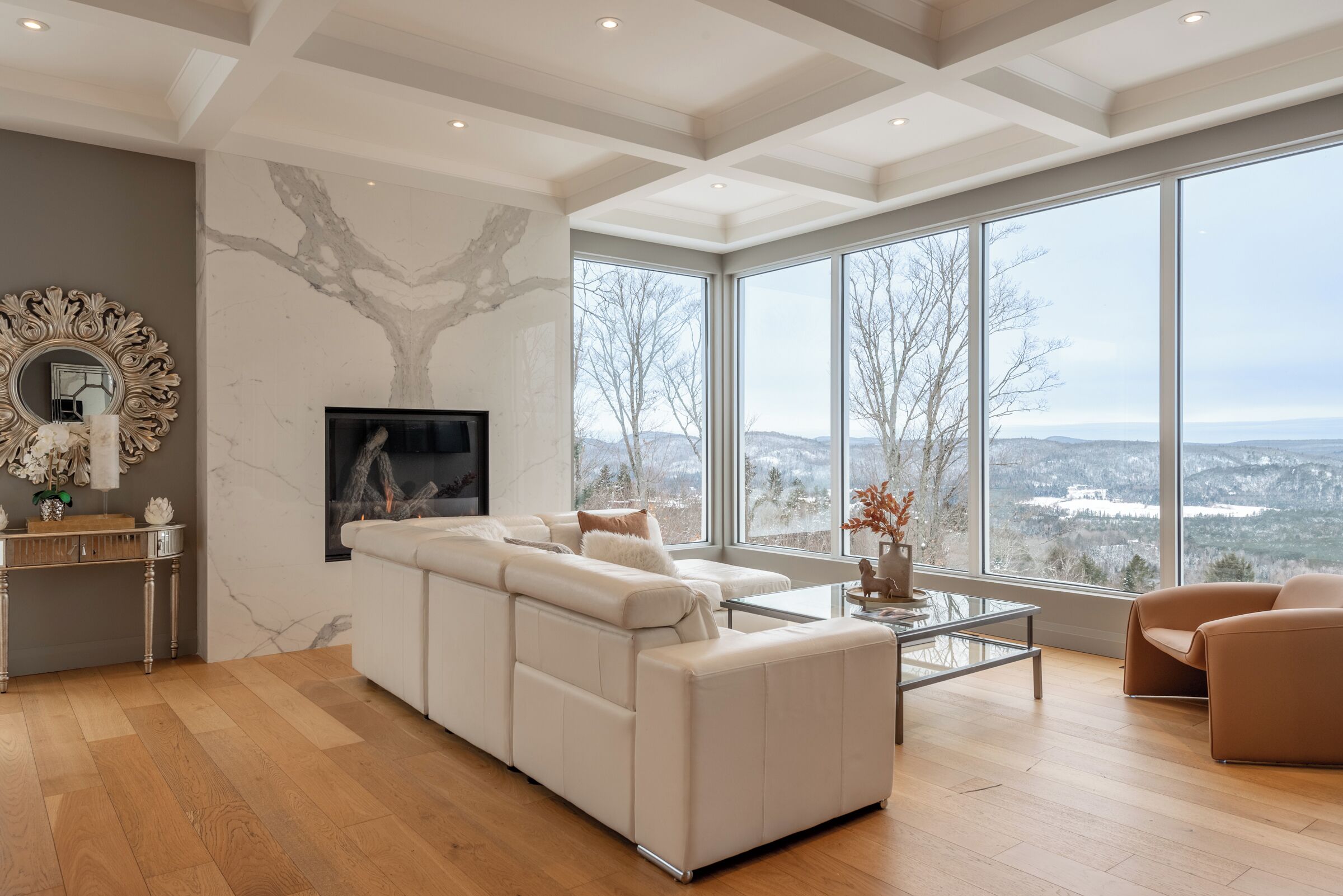 Uma sala de estar ampla com um grande sofá branco modular, uma mesa de centro de vidro e uma lareira. O espaço possui uma janela grande com vista para uma paisagem nevada.