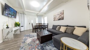 Un salon moderne avec un canapé gris, une table basse, un téléviseur fixé au mur, un coin repas avec des chaises et une table, et un tableau accroché au mur.