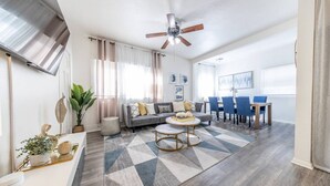 Un salon moderne avec un canapé gris, une table basse et des chaises bleues. On y trouve également un coin repas avec une table et des chaises, ainsi qu’un ventilateur de plafond.
