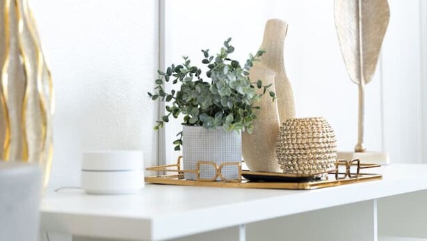 Une console blanche et moderne, avec un téléviseur fixé au mur juste au-dessus. Sur la console, on trouve un vase blanc contenant des feuilles vertes, une sculpture en bois décorative et un panier tressé.