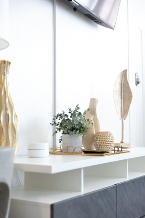 Una consola moderna y blanca con un televisor montado encima. Sobre la mesa, hay un jarrón blanco con follaje verde, una escultura de madera decorativa y una cesta tejida.
