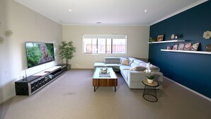 A living room with a flat-screen TV, a grey sofa, a coffee table, and a small side table with a plant. - Modern 6BRM House Big Group at Pakenham (Pakenham)