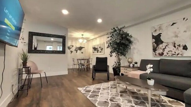 Un salon moderne avec un canapé foncé, une table basse et un tapis à motifs. On y trouve également un coin repas avec une table et des chaises, ainsi qu'une grande plante dans un coin.