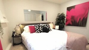 Un lit bien fait, avec une literie blanche et un plaid rose, une lampe de chevet, une petite table avec une lampe, un miroir et une plante en pot.