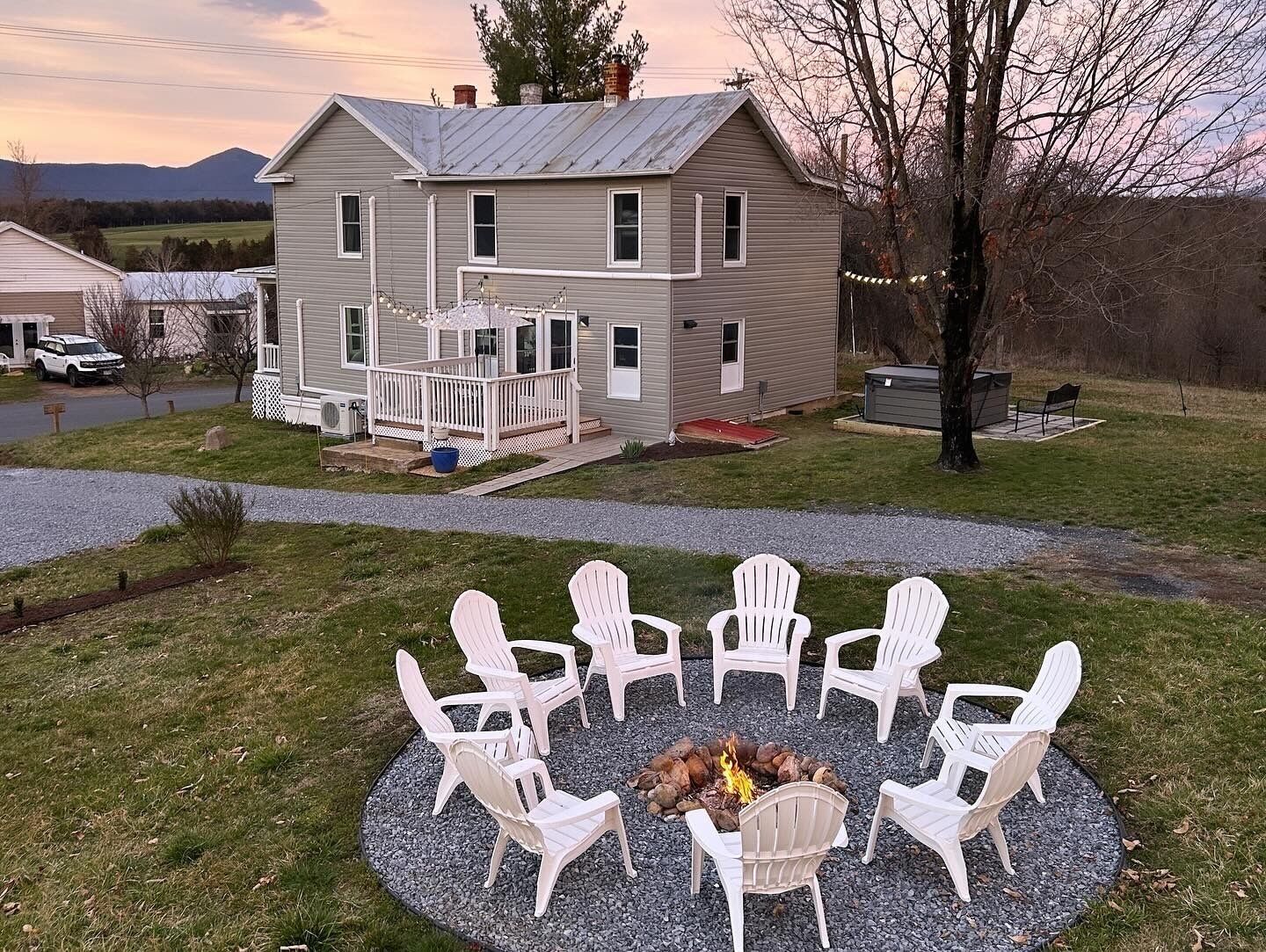 A two-story house with a porch, a fire pit surrounded by white chairs, and a gravel driveway.
