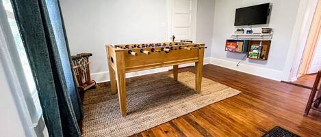 A room with a wooden foosball table, a television mounted on the wall, and a door in the background.