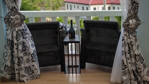 Un balcón con dos sillas de mimbre, una mesa con una botella y vasos, y cortinas con estampado.