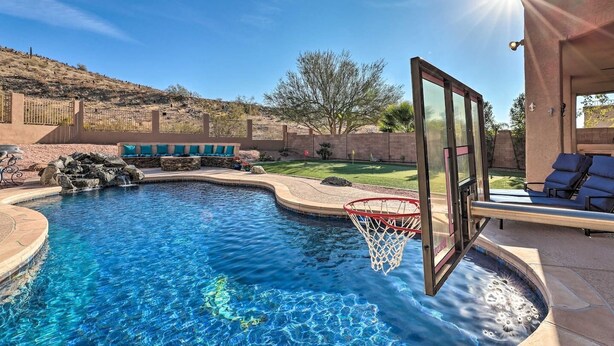 A swimming pool with a basketball hoop, a seating area with blue cushions, and a view of a hilly landscape.