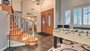 A spacious living area with a wooden staircase, a foosball table, and a large window with blinds.