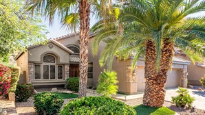 A two-story house with a stone facade, large windows, and a well-maintained garden with palm trees and shrubs.