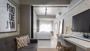 A modern living room with a grey sofa, a dining area with a white table and chairs, and a kitchen with a brick wall and a large TV. - 3 Minutes From Copacabana Beach Df115 (Rio de Janeiro)