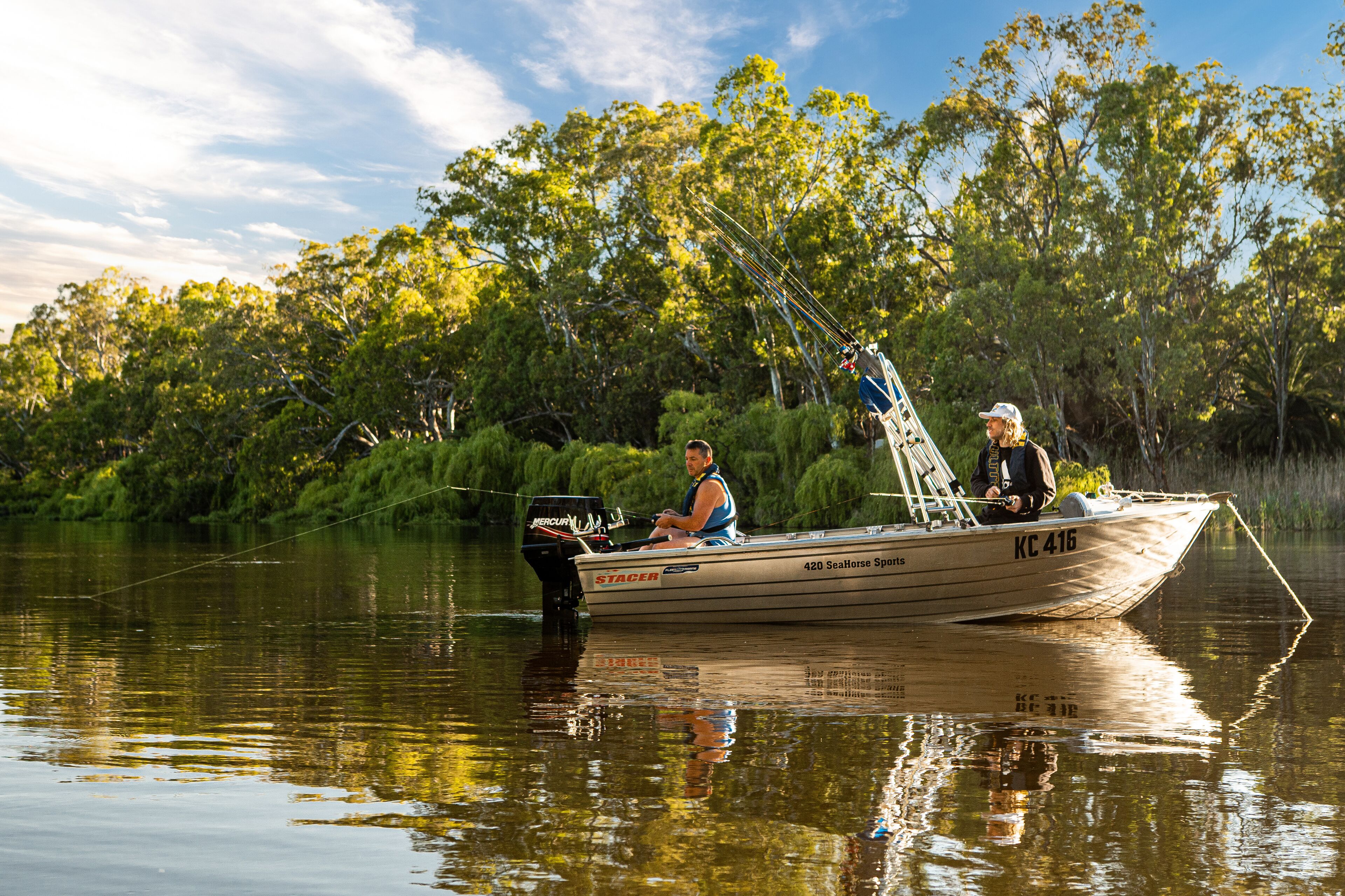 A boat with the number KC 416 on the side, equipped with a motor and fishing gear, floating on calm water with dense greenery in the background.