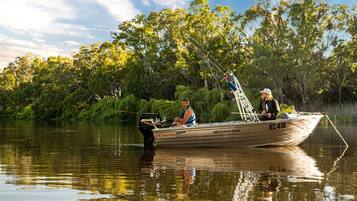 A boat with the number KC 416 on the side, equipped with a motor and fishing gear, floating on calm water with dense greenery in the background.