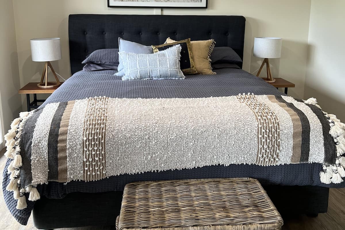 A bedroom with a large bed, two bedside tables, a wall-mounted picture, and a woven bench.
