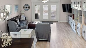 Ein geräumiges Wohnzimmer mit einer Couch, einem Hocker und einem Esstisch mit Kerzen. An der Decke befindet sich ein Ventilator, eine Uhr und ein an der Wand befestigtes Fernsehgerät.