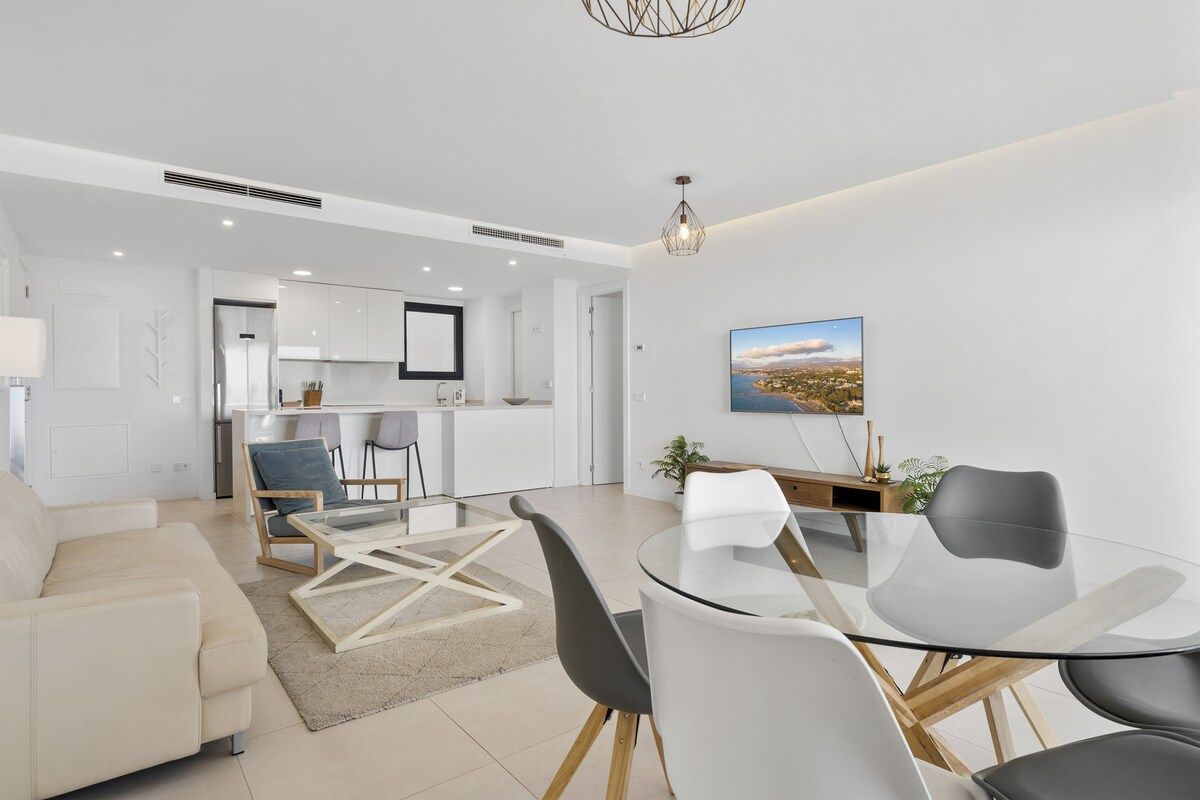 A modern living room with a glass dining table, a sofa, and a kitchen in the background.