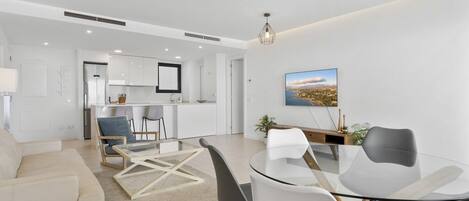 A modern living room with a glass dining table, a sofa, and a kitchen in the background.