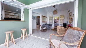 A spacious living area with a large open doorway leading to a dining area. There are two wooden stools, a wicker sofa, and a rocking chair. - Villa · St-François · Golf view & private pool (Saint-François)