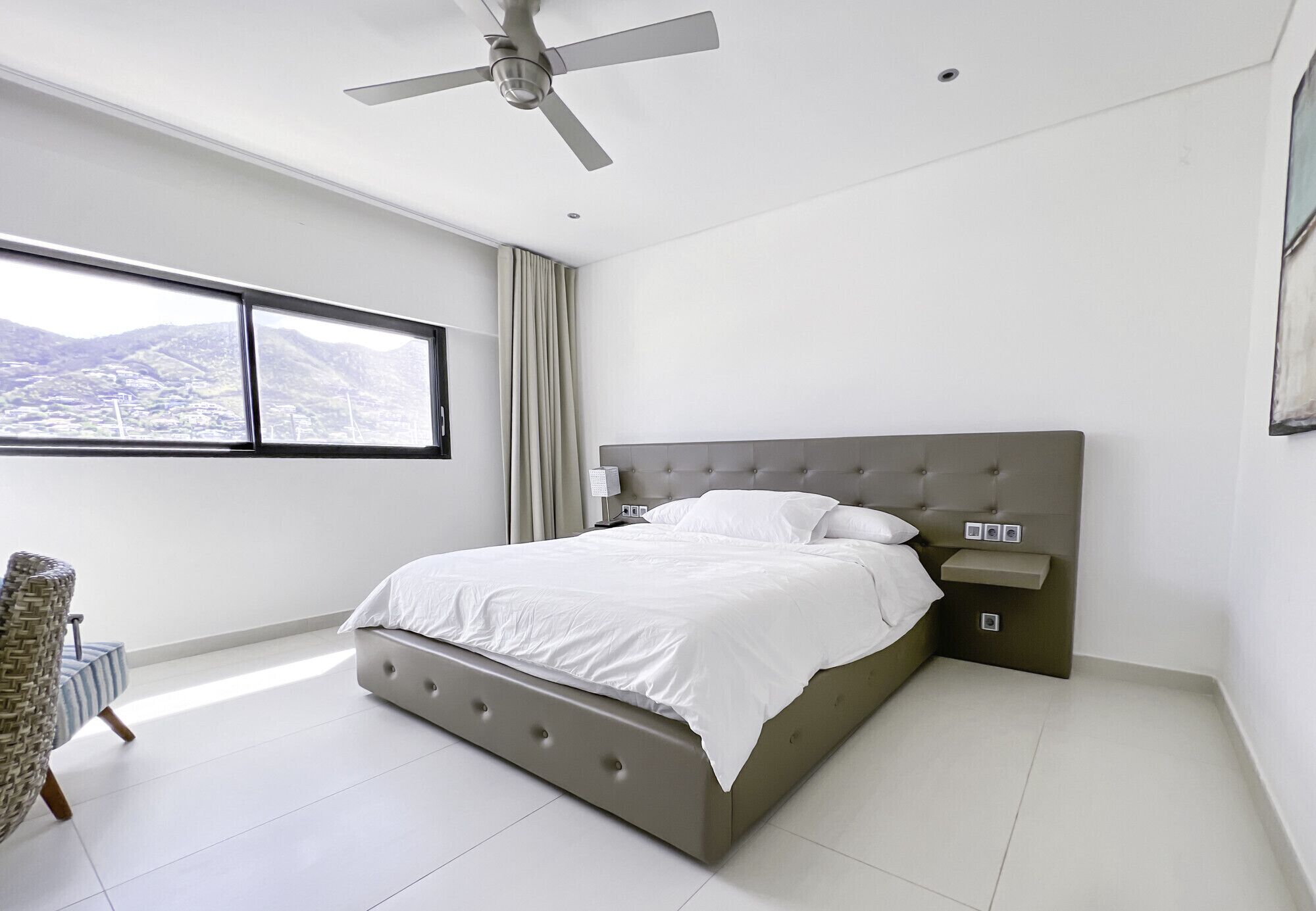 Une chambre moderne avec un grand lit, un ventilateur de plafond, une table de chevet et une vue sur les montagnes par une grande fenêtre.
