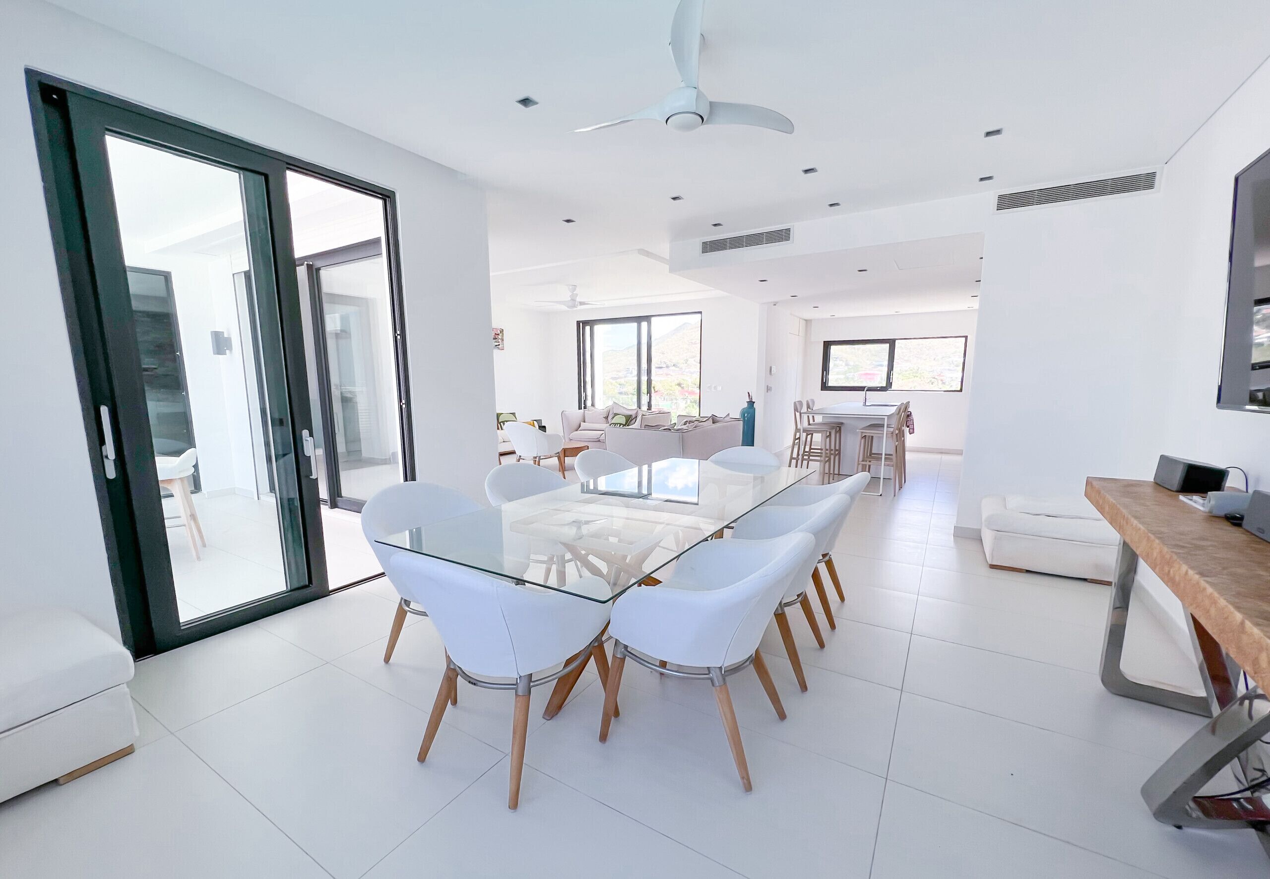 A modern, well-lit dining area with a glass-top table and white chairs, large windows, and a ceiling fan.