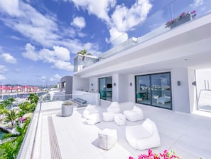 A modern rooftop terrace with white seating, large windows, and a view of the marina.