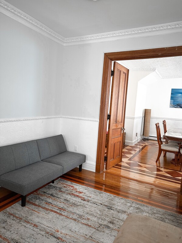 A room with a grey sofa, wooden dining table with chairs, a patterned rug, and a wooden door.