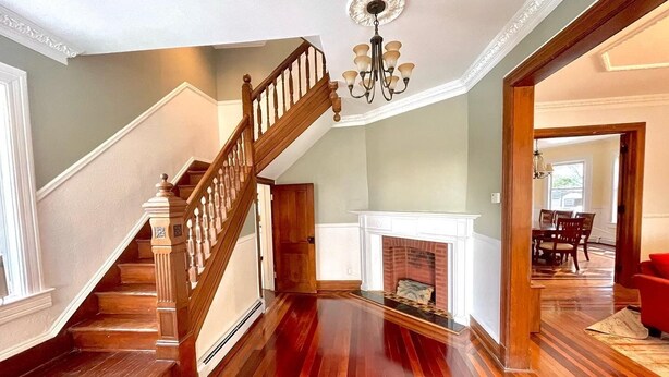 A spacious living room with a wooden staircase, a fireplace, and a dining area visible through an open doorway.