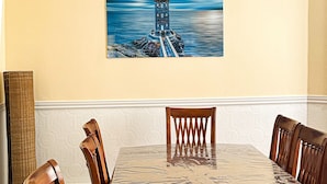 A dining room with a large table, six wooden chairs, and a painting of a lighthouse on the wall.