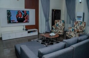 Olohuoneessa on taulutelevisio, harmaat sohvat, kuviolliset nojatuolit, sohvapöytä ja senkki.