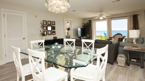 Un salon moderne avec une table à manger en verre, des chaises blanches, un lustre, un ventilateur de plafond et une vue sur l’océan.