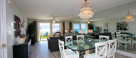 A spacious living area with a glass dining table, white chairs, a dark sofa, and a large mirror reflecting the room.