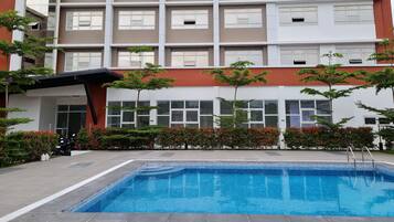 A swimming pool with tiled edges, surrounded by a low hedge and small trees. In the background, there is a multi-story building with large windows and a red and white facade.