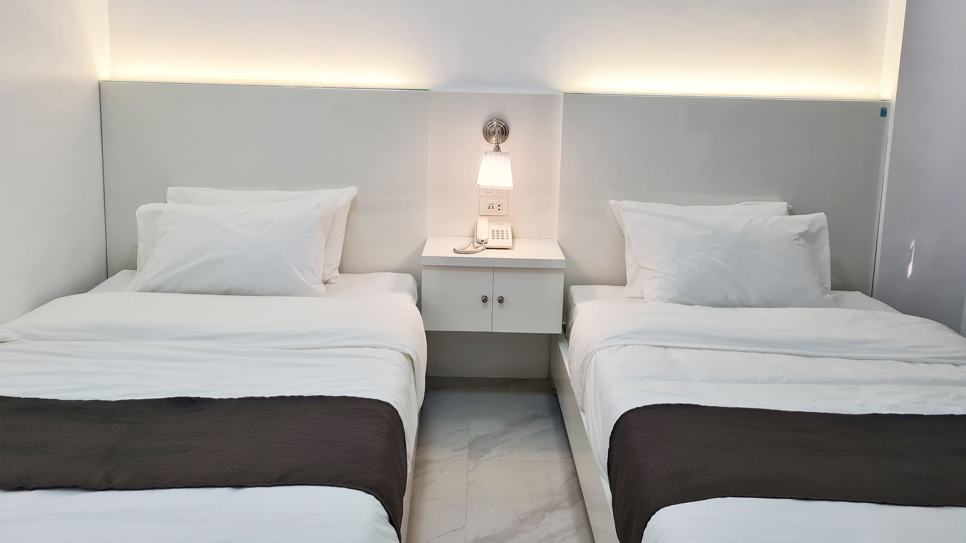 A hotel room with two beds, a bedside table with a lamp, and a small cabinet.