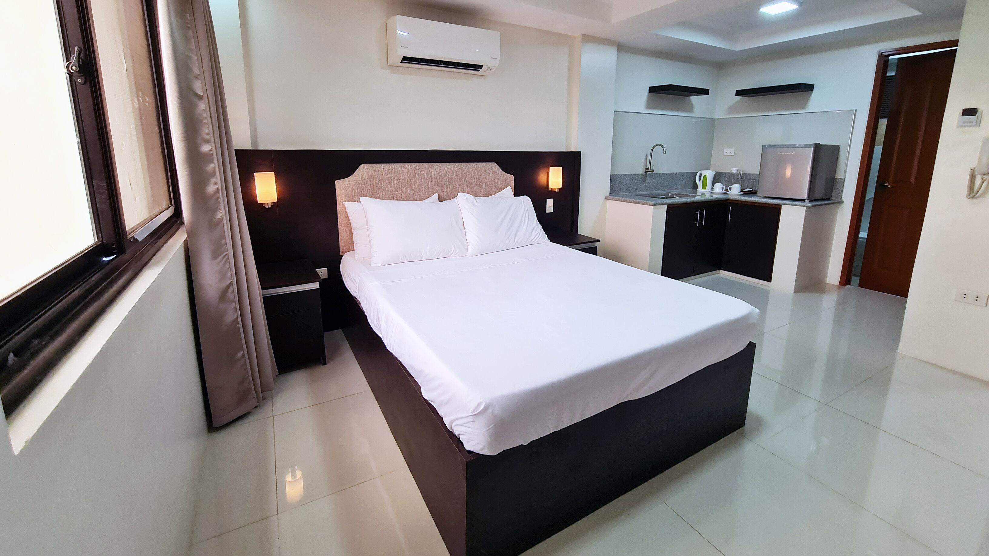 A hotel room with a bed, bedside tables, an air conditioning unit, a small kitchenette, and a bathroom.