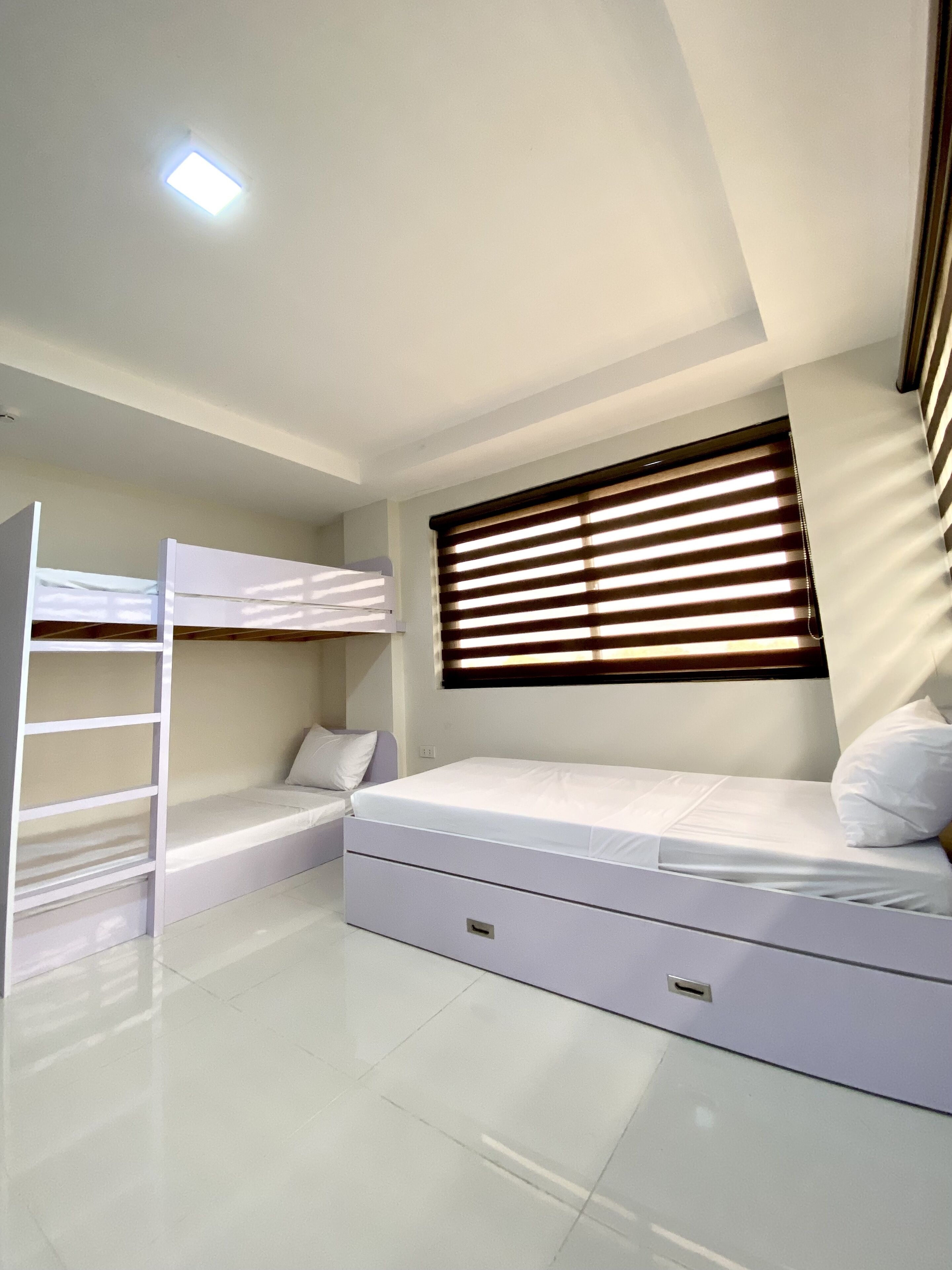 Un dormitorio con una cama litera, una cama individual, una ventana con persianas y una lámpara en el techo.