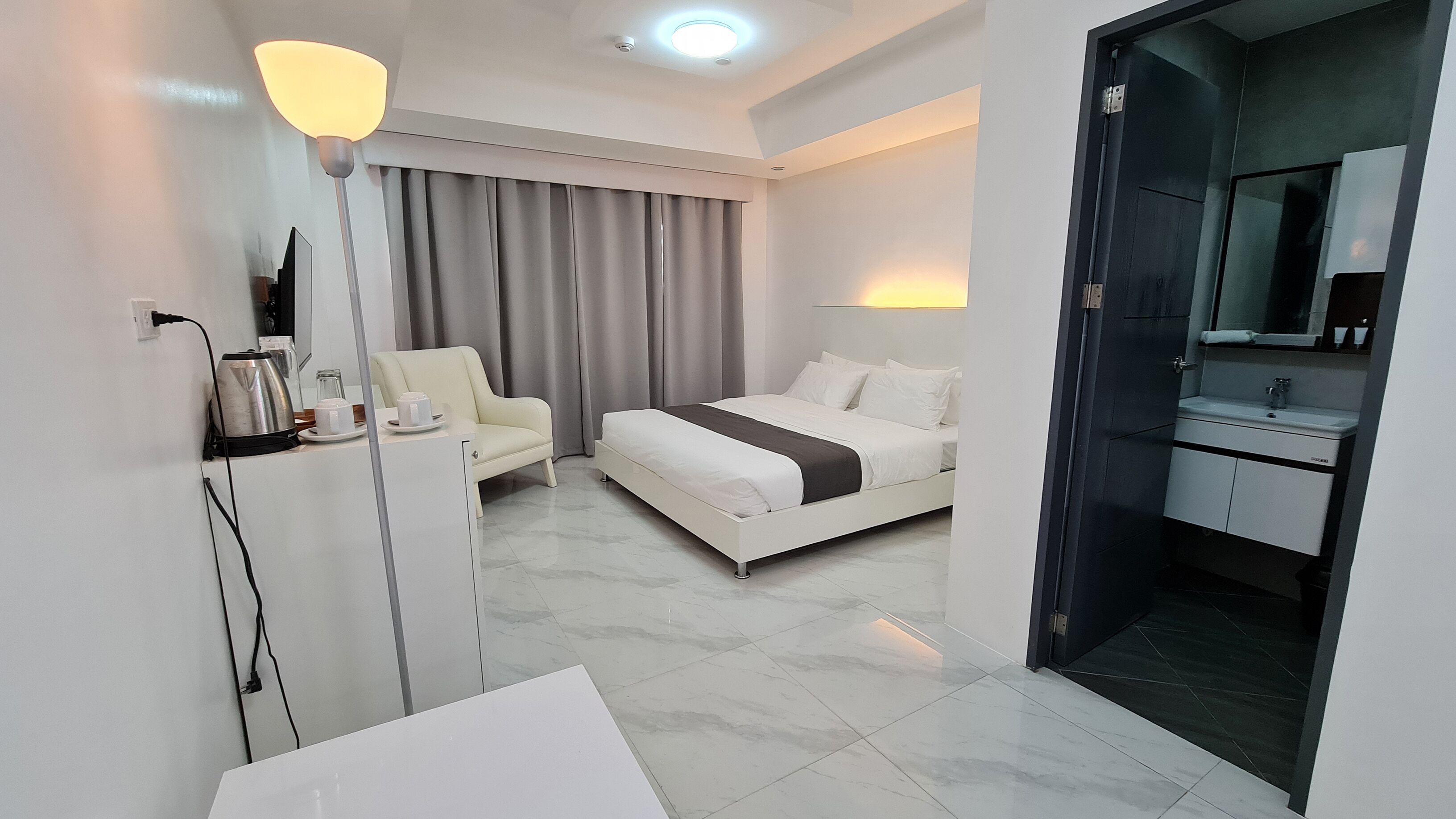 Una habitación de hotel moderna con cama, mesita de noche, lámpara y baño con espejo y lavamanos.