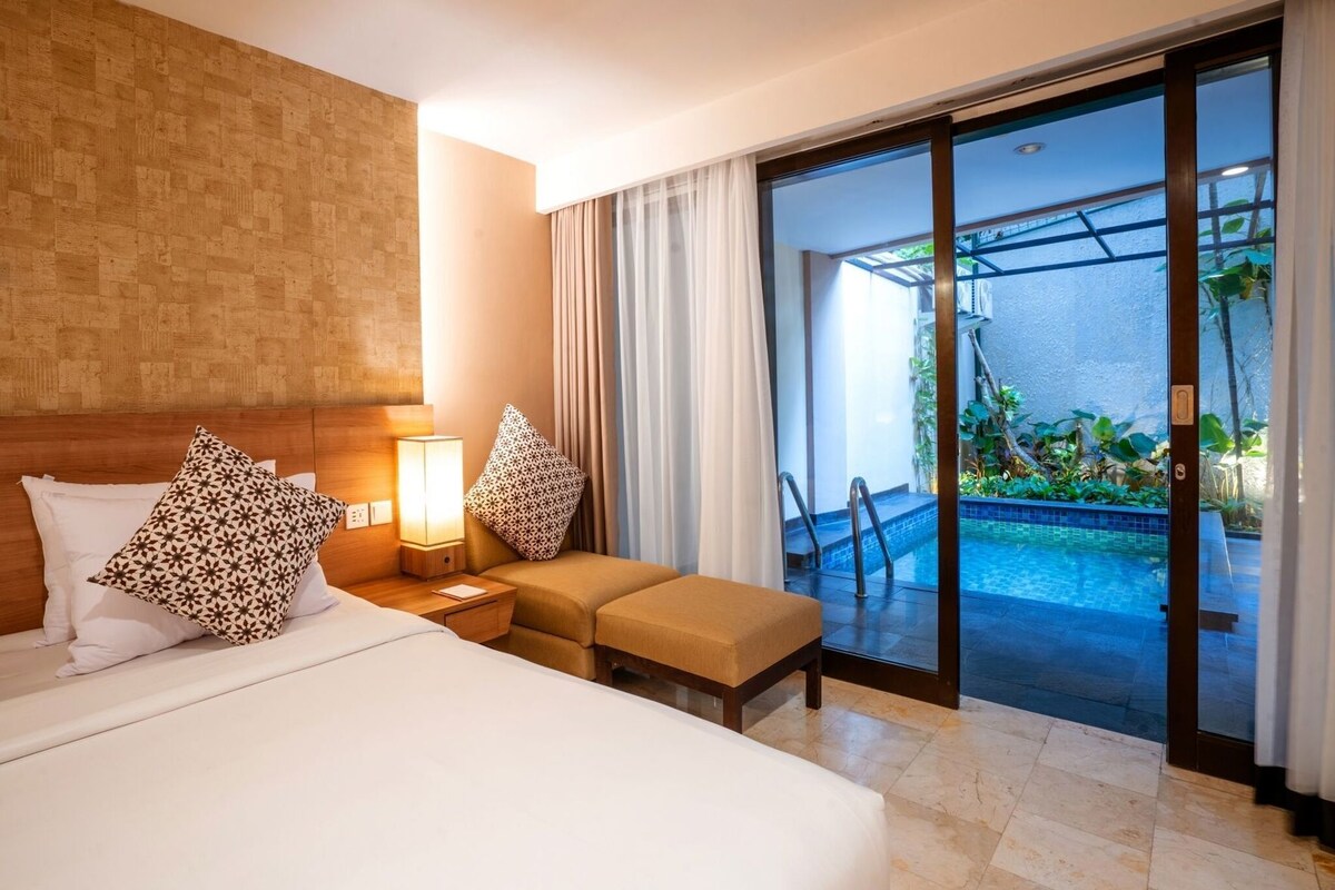 A hotel room with a bed, a nightstand, a lamp, a sofa, and a sliding glass door leading to a pool area.