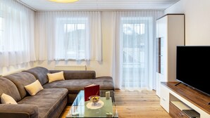 En moderne stue med sofa, sofabord, fjernsyn og store vinduer med lette gardiner.