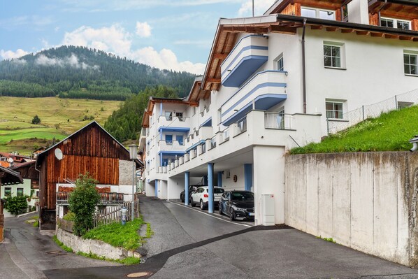 Un edificio de varios pisos con balcones, un estacionamiento y vistas a una ladera verde.