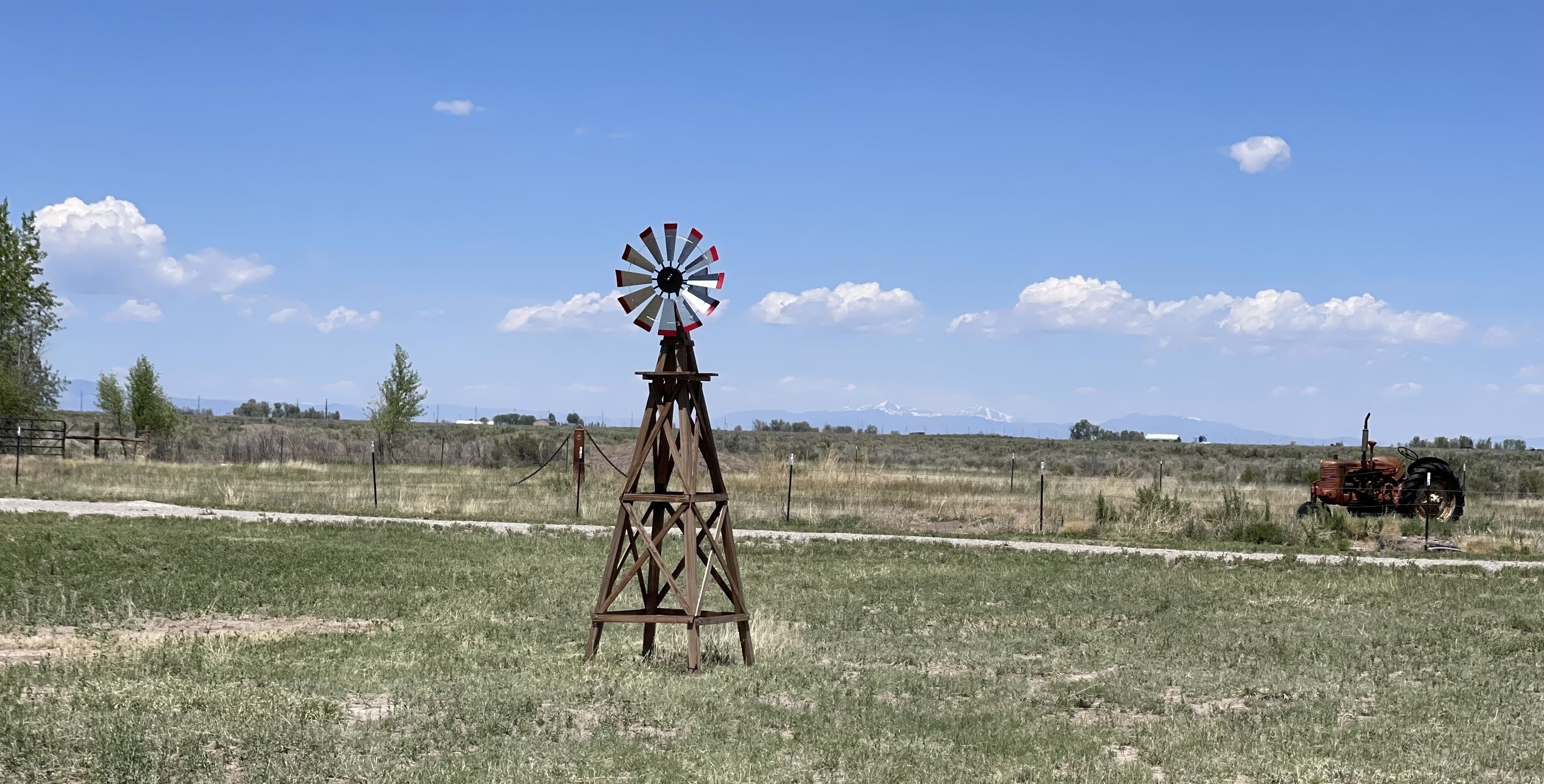 Un moulin à vent se dresse dans un champ à la campagne, avec un tracteur en arrière-plan.