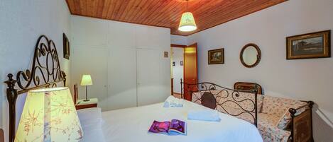 Un dormitorio con techo de madera, una cama con cubrecama blanco, una lámpara de noche y un sillón estampado con flores.