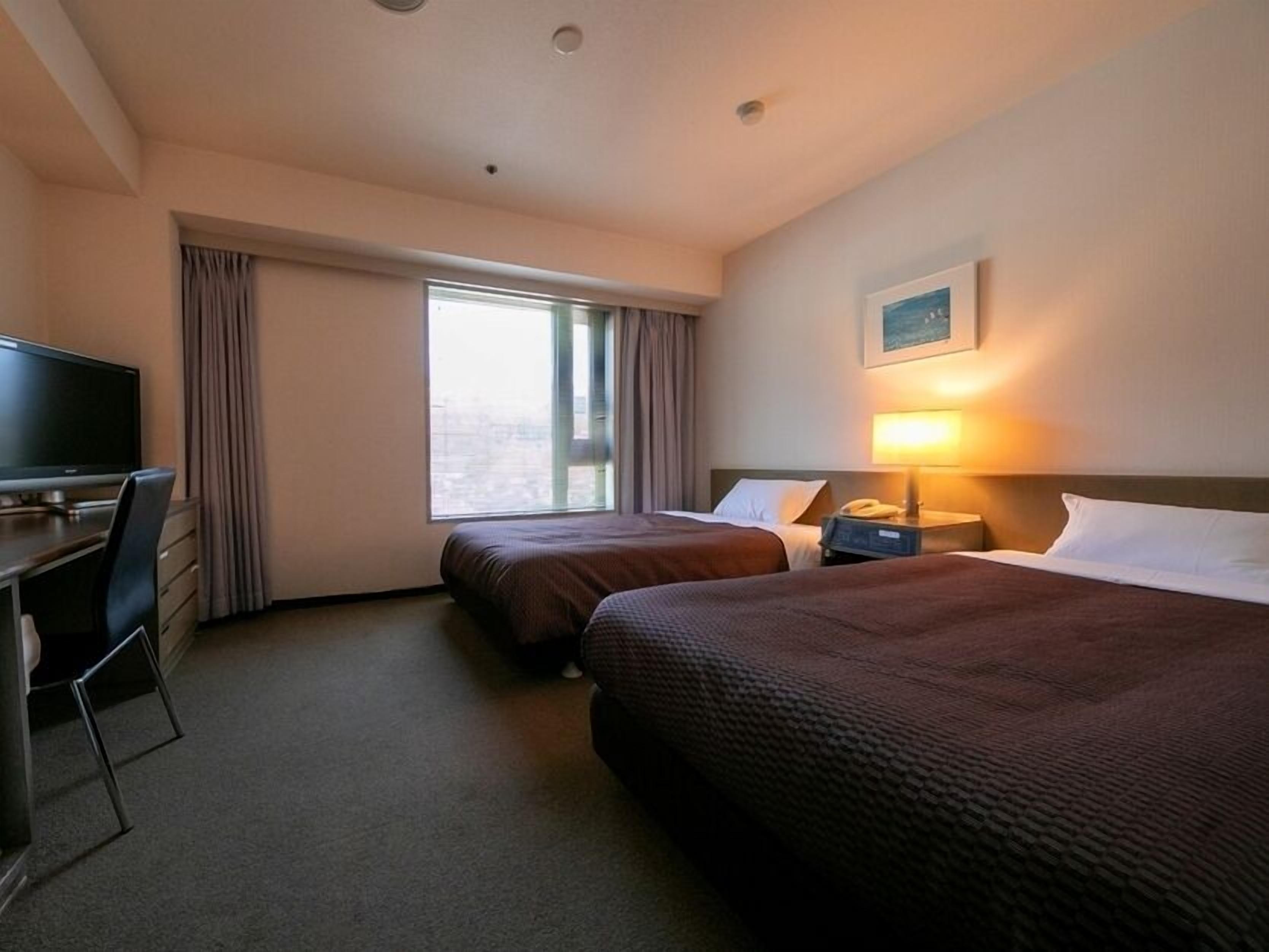 Une chambre d’hôtel avec deux lits, un bureau, une chaise, une télévision, une fenêtre avec des rideaux et un tableau au mur.