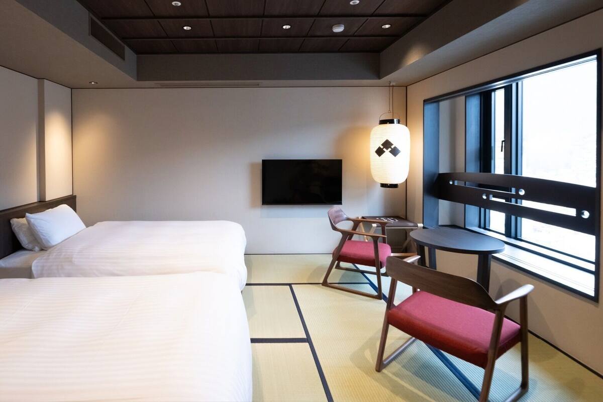Une chambre d’hôtel avec deux lits, une télévision, une table avec des chaises et une grande fenêtre.