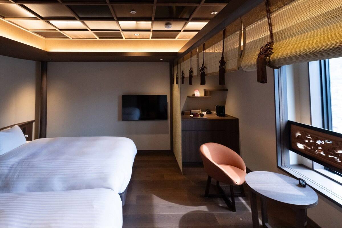 Une chambre d’hôtel avec deux lits, un bureau, une chaise, une télévision et une fenêtre avec des stores.